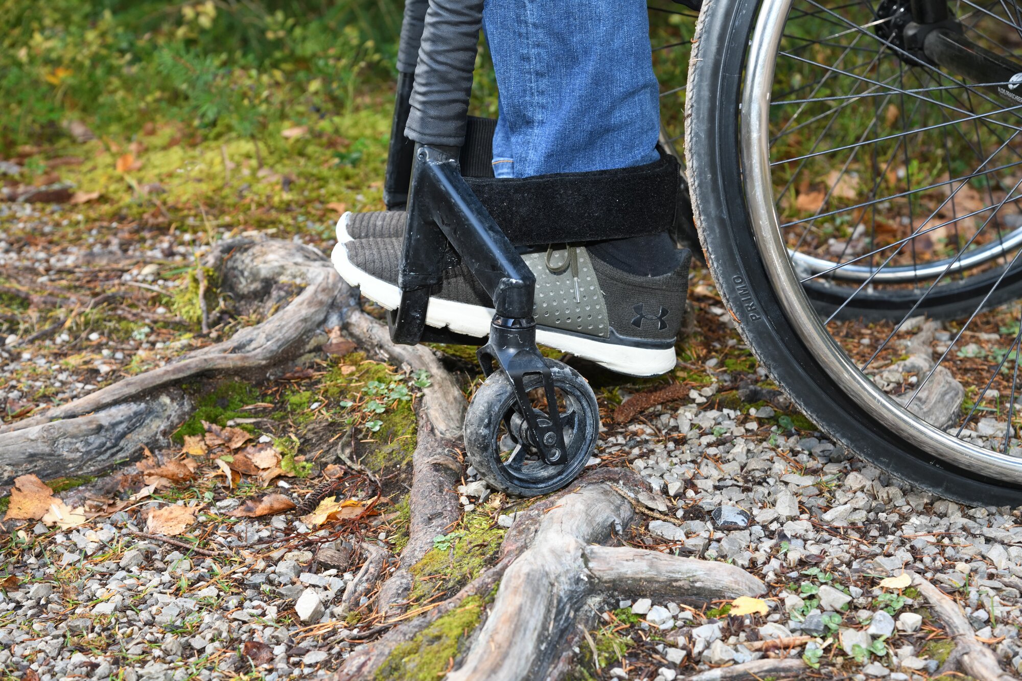 Man sieht in Nahaufnahme die Beine und Füße eines Rollstuhlfahrers und die vorderen kleinen Rollen, die an einer Wurzel anstehen.